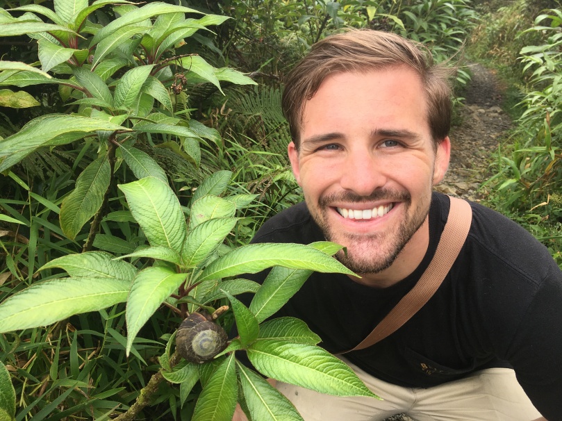 White masculine presenting person in nature smiling next to a snail